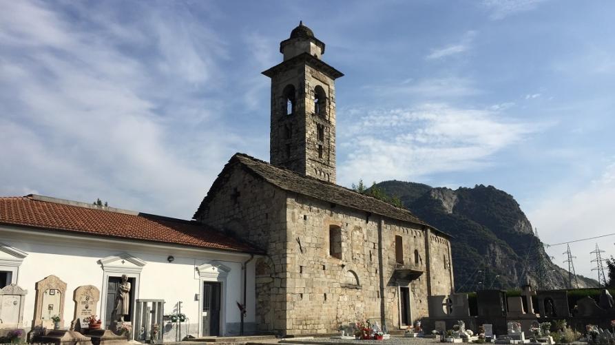 Gravellona Toce (VCO). Chiesa di San Maurizio vista laterale. La chiesa romanica in pietra grigia con campanile è fotografata dal campo santo. Il cielo è leggermente velato da nuvole bianche 