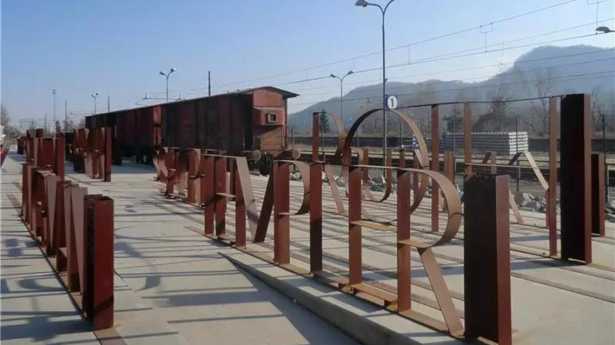Borgo San Dalmazzo (CN). Memoriale della deportazione. A destra la ferrovia e il treno fermo sui binari. In primo piano le scritte in ferro che sembrano transennare la banchina e riportano i nomi dei gruppi famigliari deportati 