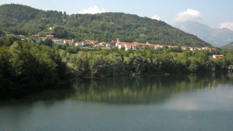 Vidracco (TO). Panorama dalla strada. In basso il fiume scorre tranquillo. Poco più in alto sul lato sinistro, tra la vegetazione, si intravede il paese sormontato da montagne ricche di alberi. Il cielo è nuvoloso 