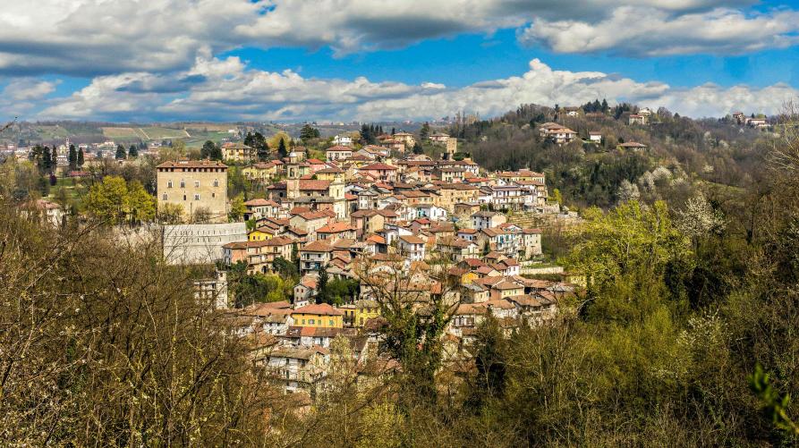 Castelletto d'Orba (AL). Panoramica del paese sparpagliato sui colli. In primo piano alberi e cespugli frondosi. Il cielo blu è solcato da nuvoloni bianchi e grigi 