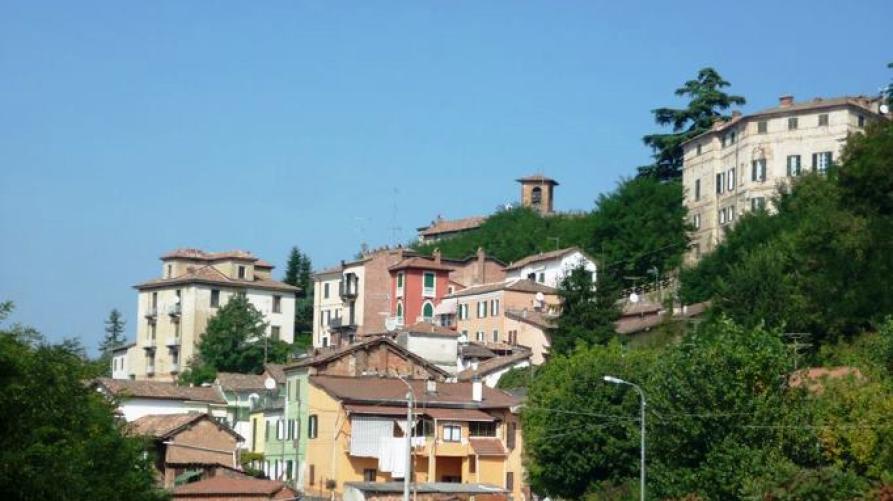 Villalvernia (AL). Le case colorate del borgo antico del paese si arrampicano a destra lungo la collina alberata. Il cielo senza nuvole è azzurro. Fotografia di Elisabetta Spotorno 