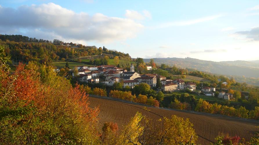 Levice (CN). In basso a sinistra alberi dai colori autunnali e un campo arato di fresco. Più in alto lo scorcio del paese che si appoggia ad una collina che degrada a destra. Il cielo azzurro è solcato da nuvole bianche e grigie. Fotografia di Annamaria Delmonte (2007)