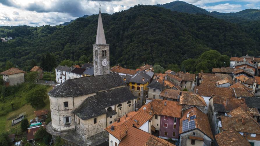 Arola (VCO). Panoramica sui tetti dalle tegole rosse del paese con vista posteriore della Chiesa di San Bartolomeo. Verso sinistra il complesso della Chiesa di San Bartolomeo vista dall'alto. Dietro al paese le montagne verdeggianti e un filo di cielo nuvoloso. Immagine tratta dal sito del Comune