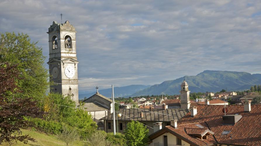 Bricherasio (TO). A sinistra prati e alberi verdeggianti da cui si intravede parte della torre parrocchiale. Sulla destra i tetti rossi del paese e in lontananza il profilo delle montagne. Il cielo è completamente coperto da nuvole grigie  