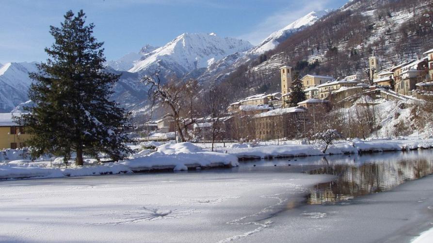 Villar Pellice (TO). Sulla sponda sinistra del fiume congelato si vede un pino frondoso e più in lontananza il paese immerso nella neve. Le montagne sullo sfondo sono completamente bianche. Il cielo è azzurro e leggermente velato dalle nuvole 