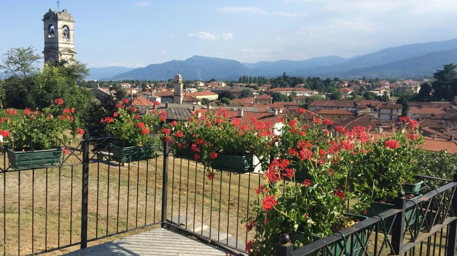 Bricherasio (TO). Vista panoramica dalla collina del belvedere. Da un terrazzino fiorito si intravedono i tetti rossi del paese e a sinistra la porzione finale del campanile della chiesa parrocchiale. Sullo sfondo le montagne e un cielo azzurro parzialmente nuvoloso 