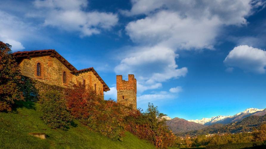 Candelo (BI). Inquadrato dal basso si vedono salire dal prato le mura, alcuni edifici e una torre in pietra viva del Ricetto medievale. Sullo sfondo le cime innevate delle montagne. Il cielo blu è solcato da nuvole bianche leggere 