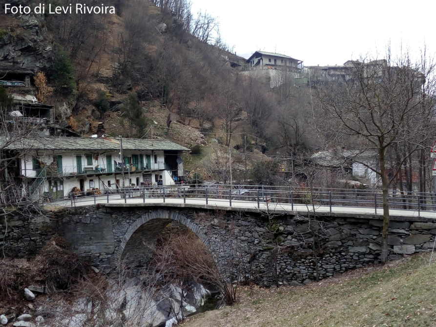 Rorà (TO). Ponte vecchio in pietre. A sinistra una cascina bianca con finestre verdi. Il bosco è stato spogliato dall'inverno. Fotografia di Levi Rivoira tratta dal sito del Comune (2018-2020) 