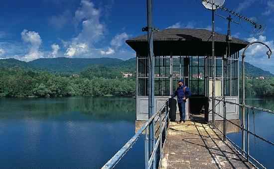 Vidracco (TO). In primo piano il camminamento in cemento della diga porta ad uno sgabbiotto. Ai lati i lago blu. In fono si intravedono gli alberi e le montagne verdi. Il cielo blu è solcato da poche nuvole leggere. Fotografia tratta dal sito del Comune (2025) 