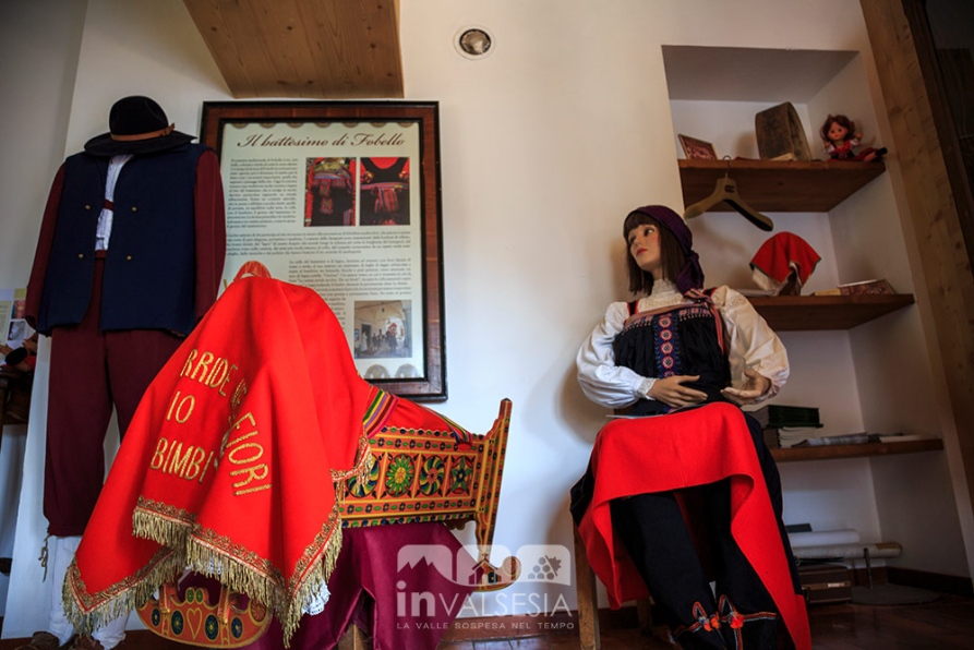 Fobello (VC). Una culla in legno dipinta di rosso e decorata con motivi verdi, gialli e blu. A sinistra un abito Walser maschile e a destra un abito Walser femminile 