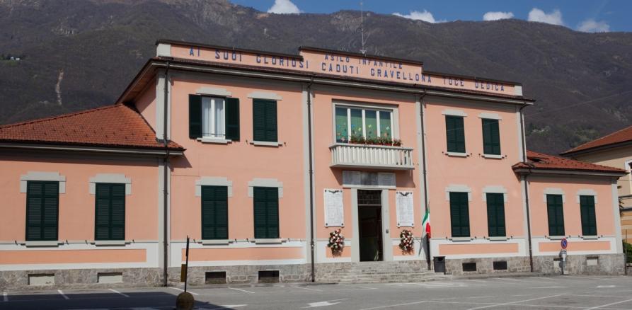 Gravellona Toce (VCO). Asilo monumento ai caduti. La costruzione a due piani con persiane verdi è dipinta di rosa. Una scritta blu appena sotto al tetto ricorda che l'edificio è sia asilo che monumento ai caduti 
