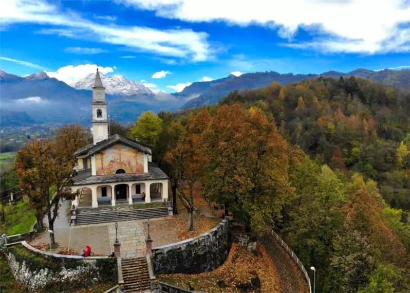 Borgo San Dalmazzo (CN). Veduta aerea del Santuario del Monserrano in basso a sinistra. A destra il bosco autunnale. In alto il cielo blu macchiato di nuvole bianche. Immagine tratta dal sito del Comune (2025) 