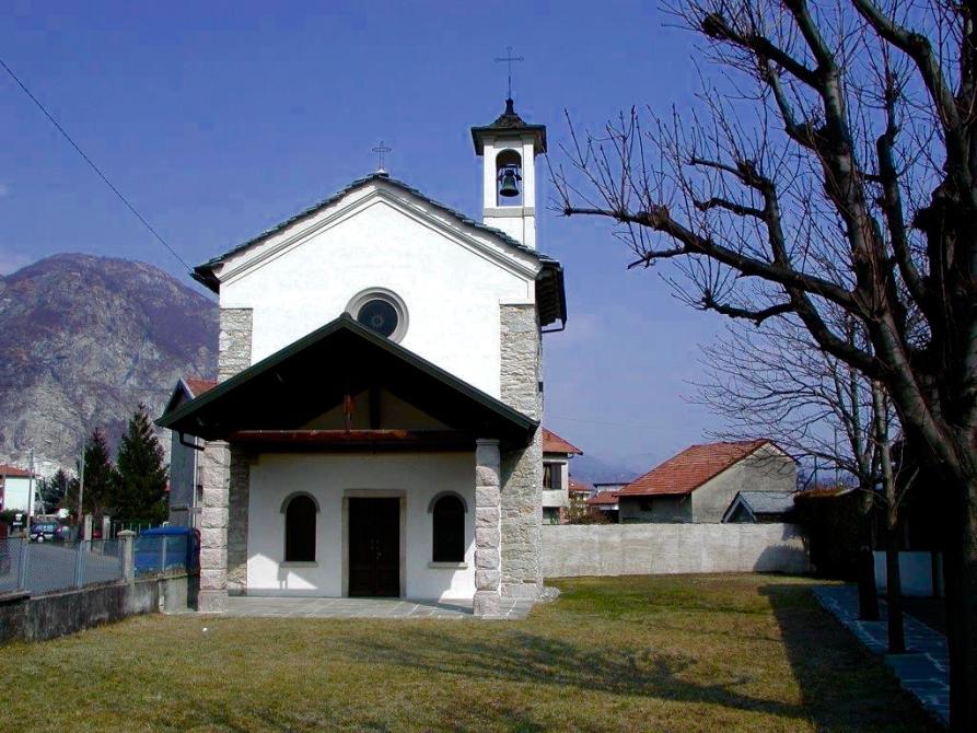 Gravellona Toce (VCO). Chiesa della Madonna dell'occhio. La chiesetta bianca con piccolo campanile poggia su un prato verde. A destra si vedono i rami spogli di alcuni alberi Il cielo è limpido. 