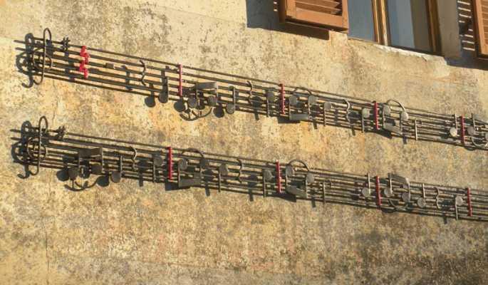 Vidracco (TO). Due righi musicali arricchiti da note in ferro scorrono su un muro di una casa. Solo il tempo e le stanghette delle battute sono colorati di rosso. Fotografia tratta dal sito del Comune (2025) 