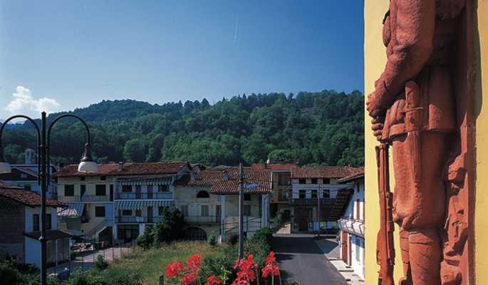 Vidracco (TO). Alcune case del paese sormontate dalla montagna ricca di alberi. Il cielo è limpido. A destra si intravede il muro giallo del municipio decorato con ceramica rossa. Fotografia tratta dal sito del Comune (2025) 
