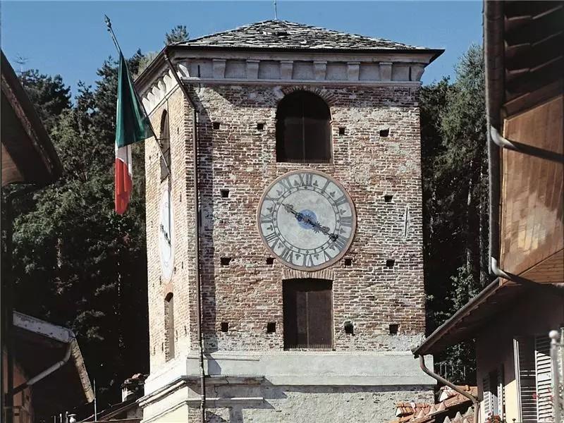 Borgo San Dalmazzo (CN). Particolare della porzione terminale della Torre civica in mattoni grezzi. L'orologio è in mezzo alle due finestre disposte verticalmente. A sinistra sventola la bandiera italiana. Immagine tratta dal sito del Comune (2025) 