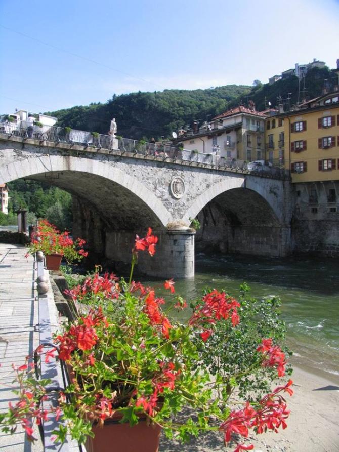 Varallo (VC). Due arcate del ponte Antonini in pietra grigia e le acque verdi del torrente Mastallone. In primo piano un vaso di geranei rossi 