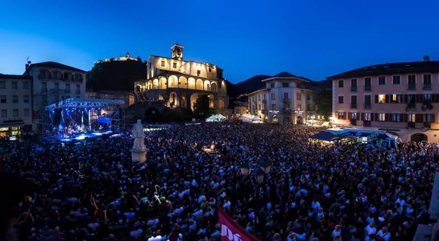 Varallo (VC). Il Sacro Monte e la Collegiata di San Gaudenzio illuminati svettano nel cielo blu elettrico. In primo piano la folla che assiste a un evento nella piazza di Varallo 