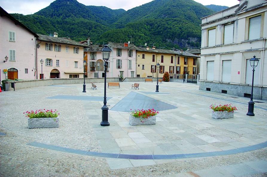 Varallo (VC). Piazzetta circolare con fioriere e panchine in legno. Le case dai muri colorati con alle spalle i monti verdeggianti. 