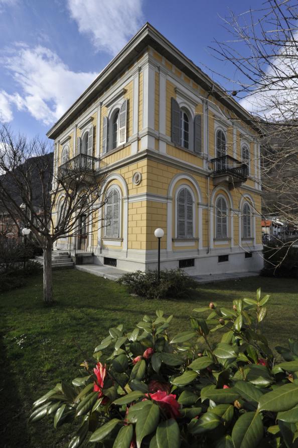 Premosello-Chiovenda (VCO). Casa Fontana-Rossi e il suo giardino. Palazzo storico a due piani con finestre ad arco e persiane grigie in risalto rispetto ai muri tinteggiati color crema 