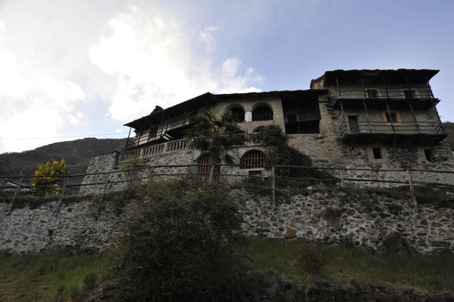 Premosello-Chiovenda (VCO). Scorcio di case arroccate in pietra e balconate in legno vecchio si stagliano su un cielo azzurro macchiato a sinistra da nuvole bianche 