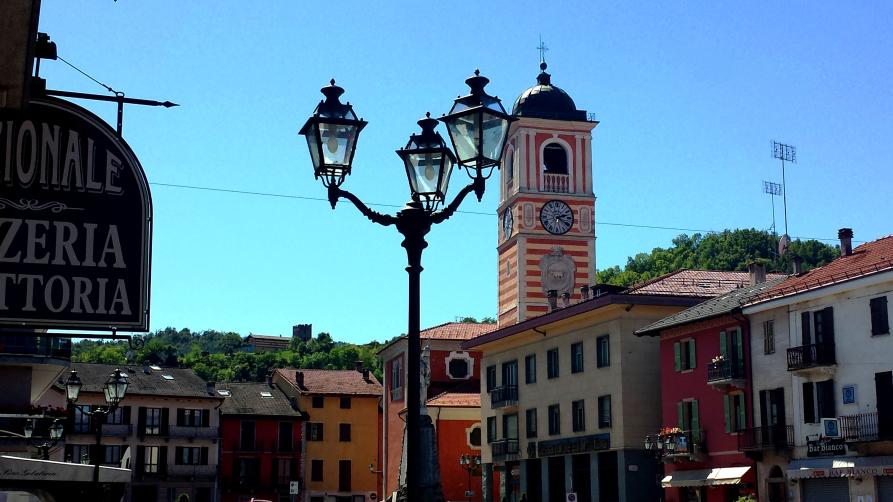 Boves (CN). Piazza Italia in primavera. Il cielo è blu. In primo piano un lampione in stile Liberty. Nel centro il campanile a righe crema e rosse. Le case coloratissime circondano la piazza. Fotografia di Giorgio Vannucchi 