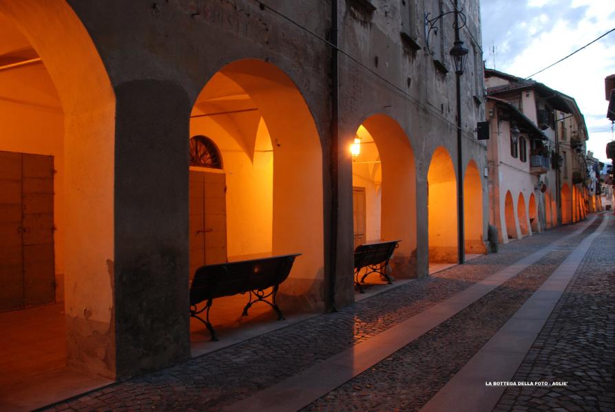 Agliè (TO). A sinistra lo scorcio dei portici del centro storico è reso accattivante dalle luci aranciate che li illuminano in contrasto con la luce pomeridiana del cielo. Fotografia di Franco Marchiando (2015). 