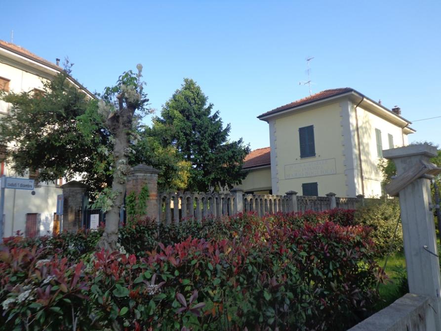 Villalvernia (AL). Una lunga siepe verde e rossiccia porta sulla sinistra all'edificio dalle finestre verdi e i muri gialli della Scuola dell'infanzia. Fotografia di Elisabetta Spotorno 