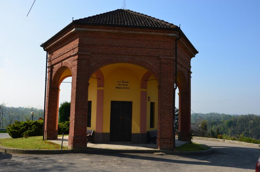 Scurzolengo (AT). Piccola Cappella di San Rocco a pianta ottogonale con porticato in mattoni rossi che la circonda integralmente. Sullo sfondo alberi e vegetazione fiorita. Il cielo è terso.   