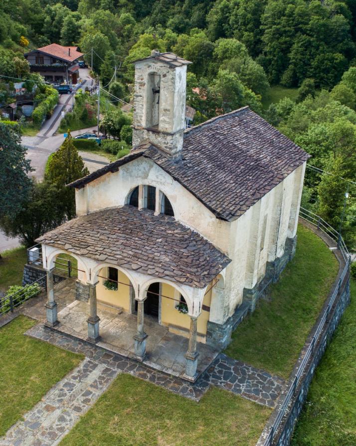 Arola (VCO). Vista dall'alto della Chiesa della Madonna dell'Assunta. Il vialetto lastricato conduce al porticato con tre archi. Svetta centrale il piccolo campanile. La copertura del tetto è in lose. Immagine tratta dal sito del Comune 