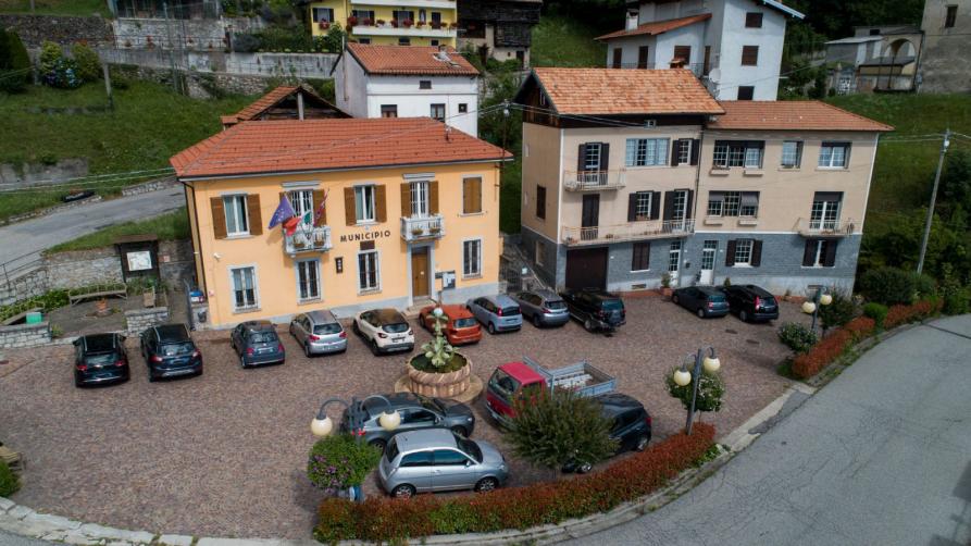 Arola (VCO). Vista dall'alto del Municipio a due piani affiancato da una casa a tre piani con persiane marroni.  Le auto parcheggiate nella piccola piazzetta abbellita da una fontana centrale. Immagine tratta dal sito del Comune 
