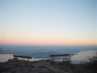 Nebbiuno (NO). Sulla rocce due panchine offrono riposo per osservare dall'alto un braccio sottile del Lago Maggiore e il territorio sottostante lontano e sbiadito. Il cielo sfuma dal color oro all'azzurro chiaro del primo crepuscolo. Immagine tratta dal sito del Comune 