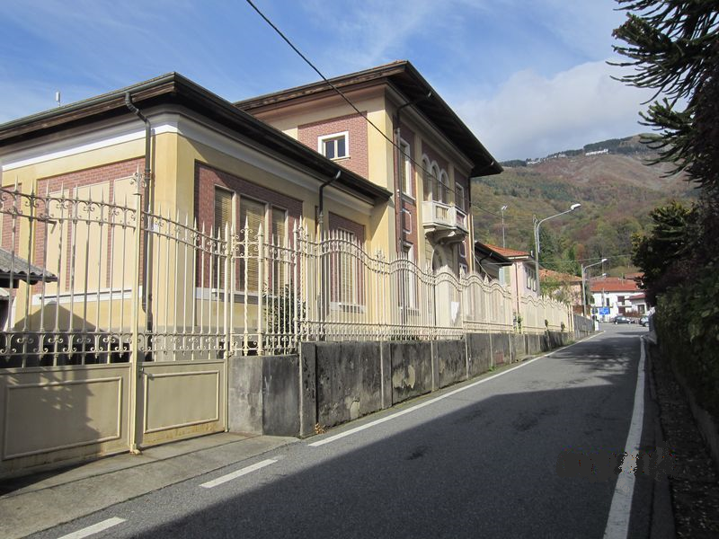 Camandona (BI). A sinistra dello stradone, delimitata da una lunga cancellata smaltata color crema, si erge la Piccola Casa della Divina Provvidenza con decori in mattoni rossi e cornici intonacate color crema. Immagine tratta dal sito del Comune (2024) 