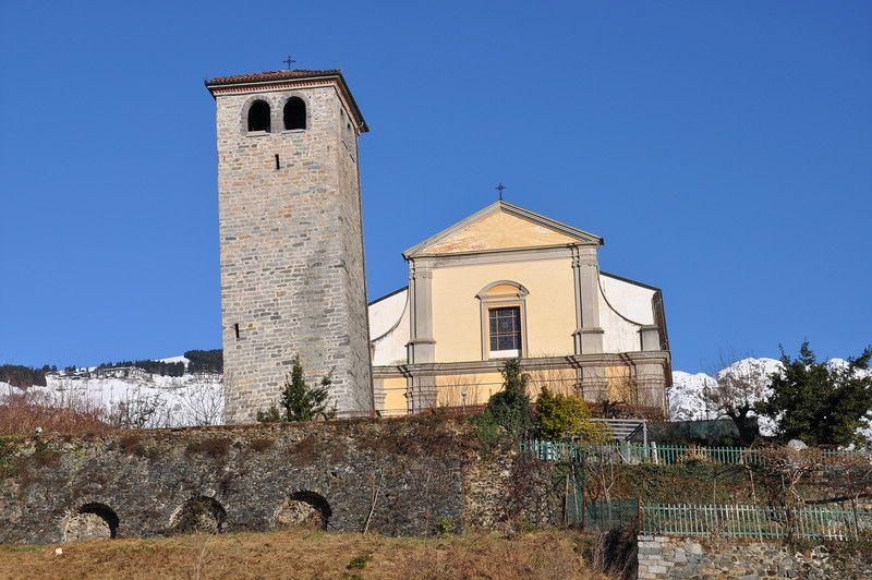 Camandona (BI). La Chiesa parrocchiale intonacata color crema e a sinistra il campanile pendente in pietre grigie hanno alle spalle le montagne innevate e un cielo blu intenso. Fotografia di Ilario Guelpa Piazza 