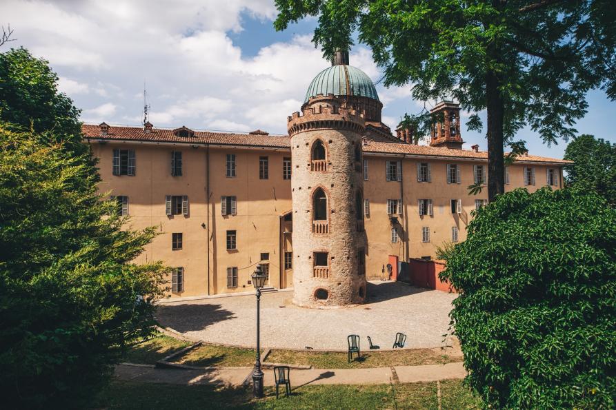 Dogliani (CN). L'edificio finestrato del municipio è intonacato color terra ed è inquadrato fra alberi frondosi. Al centro della foto una torre antica merlata a tre piani. Immagine tratta dal sito del Comune (2025) 