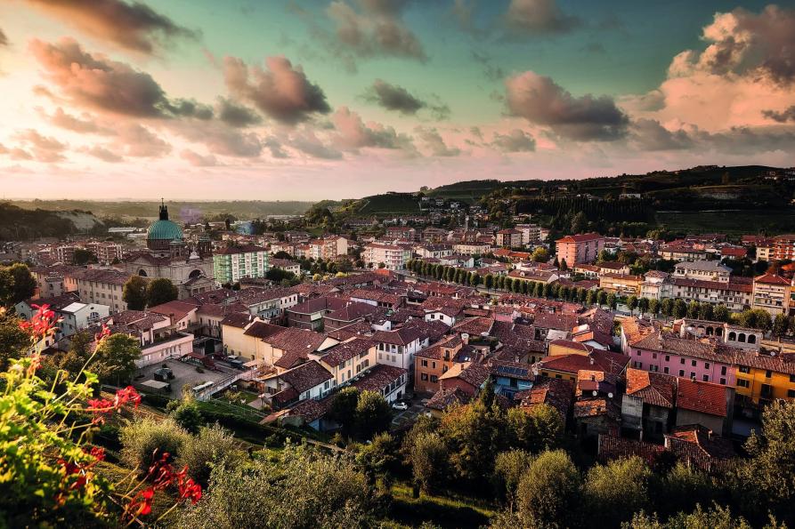 Dogliani (CN). Il paese, visto dall'alto si dipana fino alle colline verdeggianti a destra. Il cielo rosaceo e azzurro è solcato da nuvole. Immagine tratta dal sito del Comune (2025) 