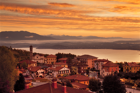 Nebbiuno (NO). Il paese al tramonto è completamente tinto di arancione. Si vedono uno specchio di lago in lontananza e le montagne violacee in pieno contrasto con i colori dorati del cielo e delle nuvole che lo solcano. Immagine di Simone Zanni tratta dal sito del Comune 