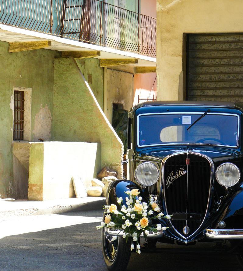 Lozzolo (VC). A destra il muso di una macchina Balilla nera è adornato di fiori bianchi. L'auto è parcheggiata all'ombra fra le mura di case di cortile 