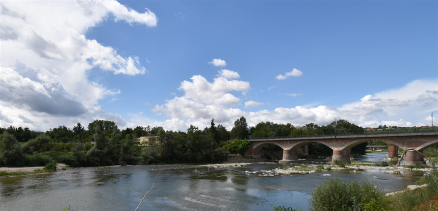 Farigliano (CN). Il fiume Tanaro scorre sotto al ponte dalle ampie arcate in mattoni rossi e grigi. Il cielo blu è solcato da ciuffi di nuvole bianche 
