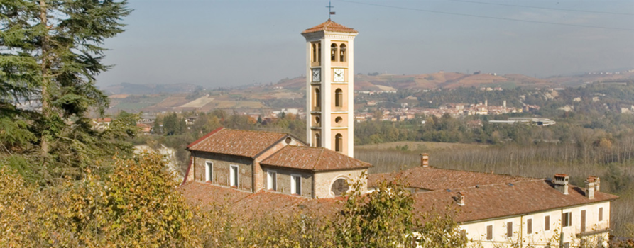 Farigliano CN). Dalla vegetazione autunnale emergono i tetti del complesso del Santuario di Nostra Signora delle Grazie di Mellea e l'alto campanile con monofore e bifore. Il cielo è velato da leggera foschia. Immagine tratta dal sito del Comune (2025) 