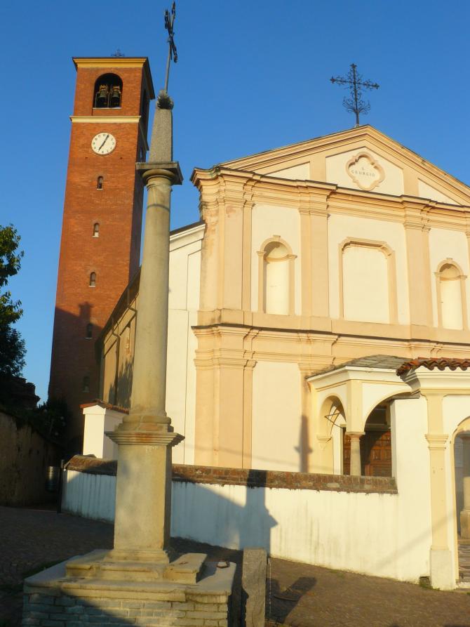 Lozzolo (VC). In primo piano una croce su colonna. A sinistra il campanile in mattoni rossi e al suo fianco la facciata barocca della Chiesa di San Giorgio 