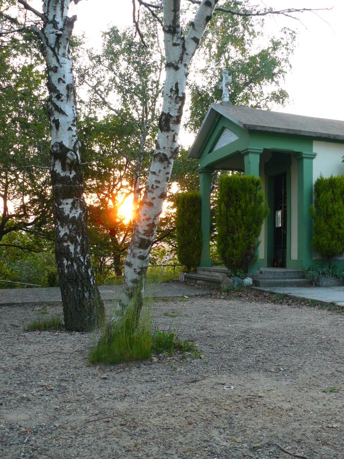Lozzolo (VC). Fra due alti alberi il sole calante al tramonto. A destra la Chiesetta degli Alpini dipinta di verde 