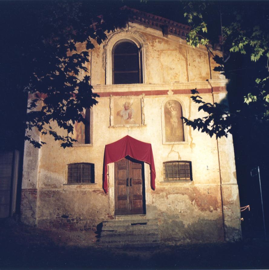 Momo (NO). Immagine notturna della facciata illuminata di San Rocco. Il portale di ingresso è sormontato da un drappo rosso. A lato due finestre con inferriate. Al di sopra tre decorazioni affrescate in due nicchie laterali e in una cornice centrale. In alto un'ampia finestra ad arco. Fotografia di Luciano Amaranto (1985) 