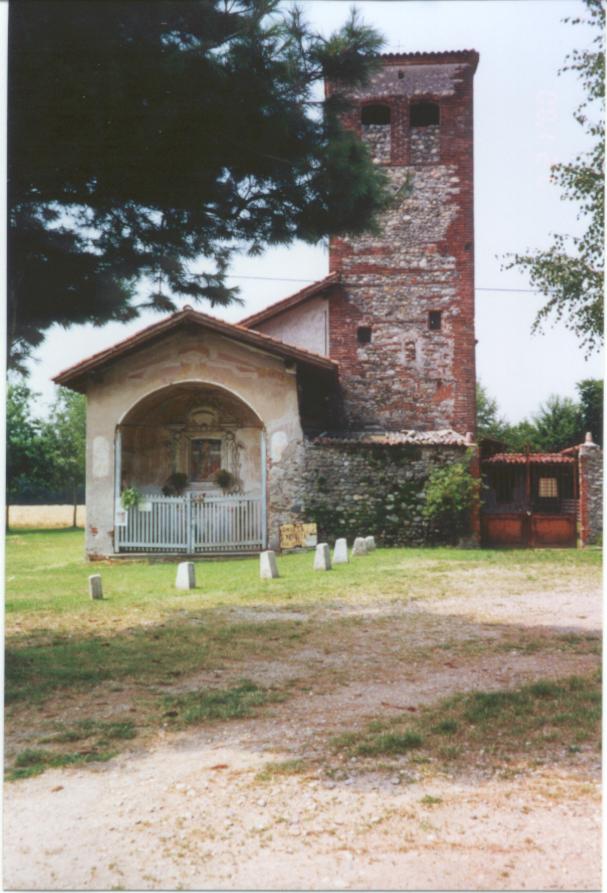 Momo (NO). Dal prato sterrato si giunge all'entrata della Chiesa della SS. Trinità. Lo spesso campanile in mattoni e pietre affianca a destra la piccola cappella interamente affrescata. Fotografia di Luciano Amaranto (2000) 