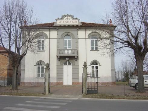 Gamalero (AL). La facciata dell'asilo "Carlo Bado" . Edificio a due piani tinteggiato di bianco con cornici grigie. Ai lati due alberi spogli. 