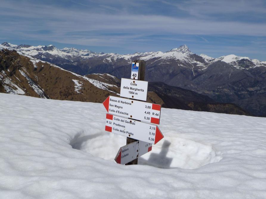 Castelmagno (CN). Colle Margherita. La neve è alta e copre il palo della segnaletica dei percorsi escursionistici. Sullo sfondo le montagne brulle spruzzate di neve e il cielo coperto da nuvole bianche sfilacciate. Fotografia di Dario Donadio 