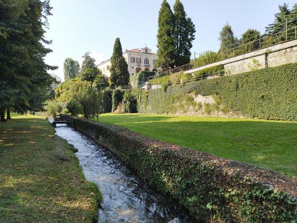 Bricherasio (TO). Un ruscello scorre al centro della fotografia e attraversa un parco con alberi e prati ben curati. A destra un muraglione ricoperto di edere verdi e in alto, fra gli alberi, si intravede la facciata del palazzo dei Conti di Bricherasio 