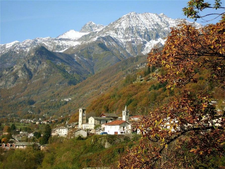 Villar Pellice (TO). A destra le fronde rossastre di un albero coprono leggermente la visuale in lontananza del paese. La vegetazione è autunnale. Sullo sfondo le montagne con le cime leggermente imbiancate. Il cielo è azzurro 