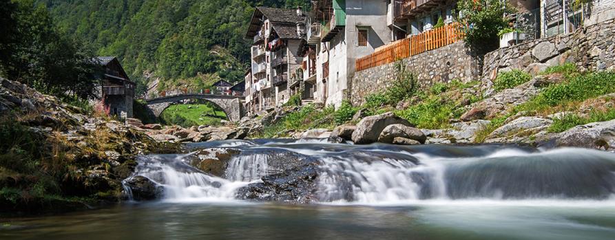Rassa (VC). Vista del paese dal torrente. Sulla riva destra le case. In fondo il ponte antico. Immagine tratta dal sito del Comune (2023) 