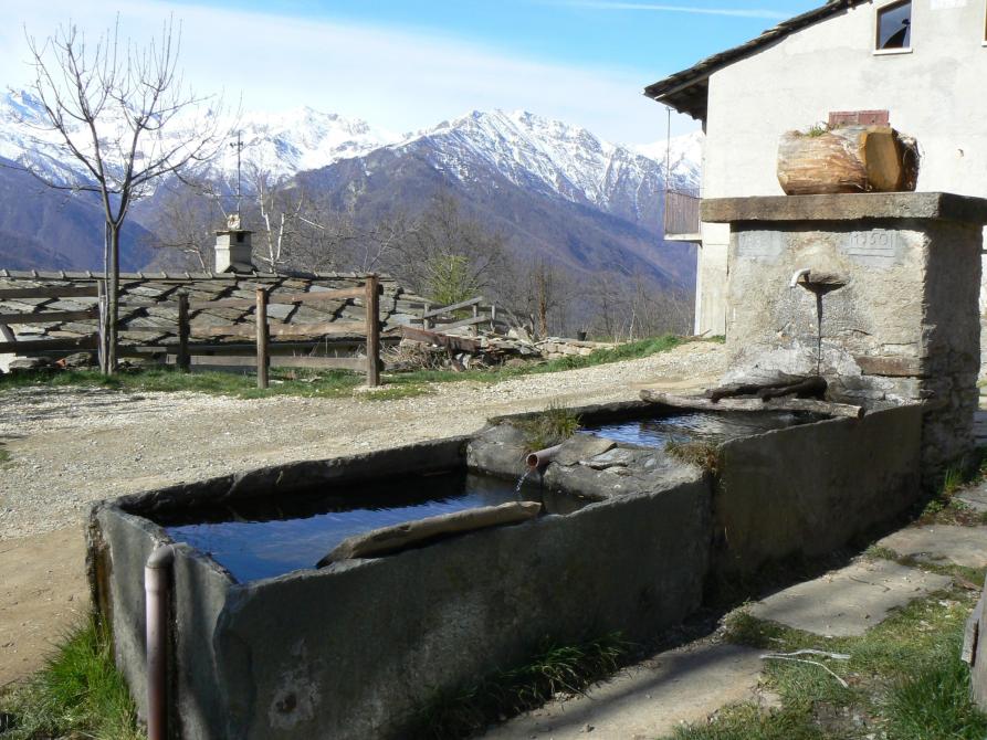 Villar Pellice (TO). A destra una tipica fontana in pietra con doppia vasca di una borgata del paese. Sullo sfondo un tetto coperto da lose e le montagne innevate 